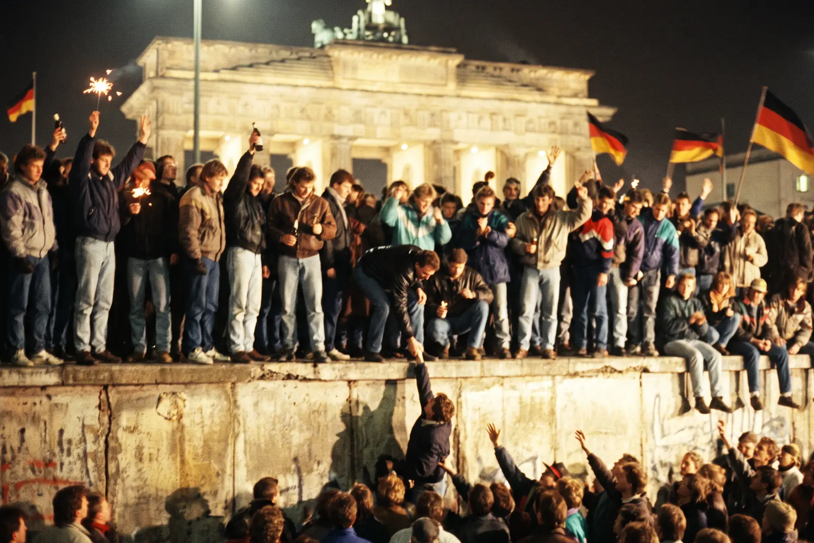 Menschen stehen und tanzen nachts auf der Mauer vor dem angestrahlten Brandenburger Tor, einige schwenken Deutschlandfahnen